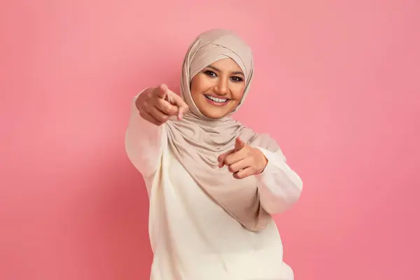 阿拉伯妇女头戴头巾 双手对着相机积极地指指点点 年轻的穆斯林女子在粉红的工作室背景上摆出一副笑脸 象征着互动和文化震动 复制空间 — 图库照片
