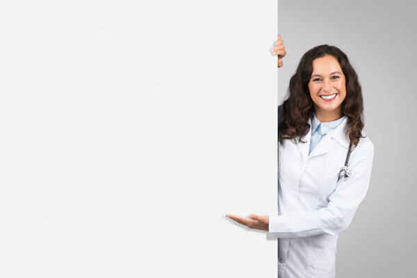 Cheerful female doctor with stethoscope peeking out from behind blank white board, presenting space for text or graphics, grey background