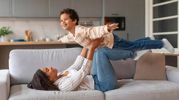 Laughing Black Mother Lying Couch Lifting Her Son Airplane Game — Stock Photo, Image