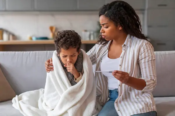 Worried black mother holding thermometer and looking at her coughing son wrapped in blanket, african american mom taking care about male child during sickness, sitting on couch in home environment