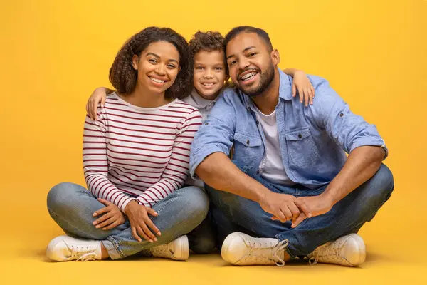 3人の幸せなアフリカ系アメリカ人の家族が床に座って抱擁し 陽気で愛情あふれる黒人の両親が黄色いスタジオの背景に息子と一緒にポーズし カメラを見て スペースをコピーします — ストック写真