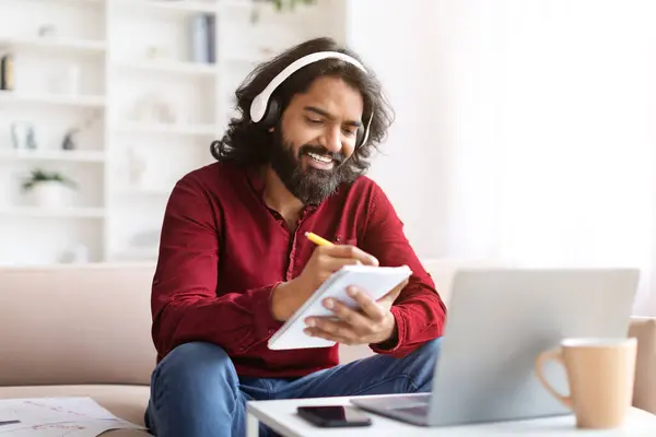 E教育について 楽しいミレニアル世代のひげ長髪のインドの男は 家庭でオンライントレーニングを持っています 居心地の良いリビングルームのインテリアでソファに座って ノートパソコンとワイヤレスヘッドフォンを使用して ノートパッドでメモを取る — ストック写真