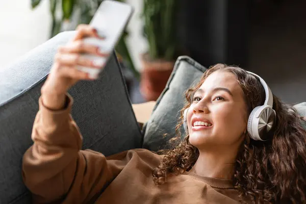 European teen girl in wireless headphones using smartphone, enjoying her weekend with music and social media, making selfie, lying relaxed on couch at home interior. Gadgets, technology
