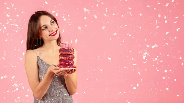 笑顔のミレニアル世代の慎重な女性は ピンクの背景で祝う菓子のフレリーの中で誕生日のろうそくで飾られたピンクのドーナツの積み重ねを賞賛する輝くドレスで — ストック写真