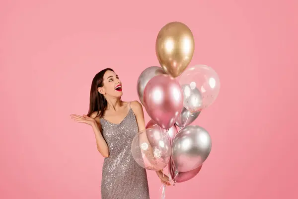 Enthusiastic caucasian millennial woman in a glittering dress laughing and gesturing with a hand as she holds a bouquet of metallic and clear balloons, with a festive pink backdrop