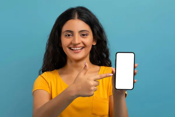 兴奋的年轻东部女学生穿着黄色的T恤 指着有白色空白屏幕的大手机 展示手机应用程序 在线服务 蓝色背景 复制空间 — 图库照片
