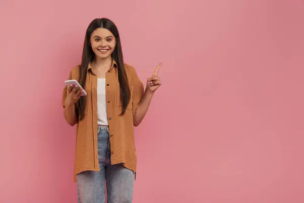 快乐的少女手持智能手机 一只手指向上 微笑的少女表示好主意或促销 站在粉色工作室的背景下 复制空间 — 图库照片