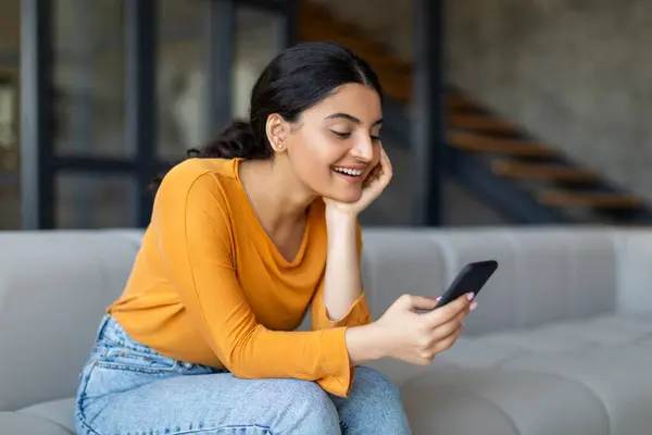 自宅のリビングルームで彼女のスマートフォンを使用して幸せな若いインドの女性 快適にソファーでラウンジング 携帯電話でインターネットを閲覧したり 友人とのメッセージング — ストック写真