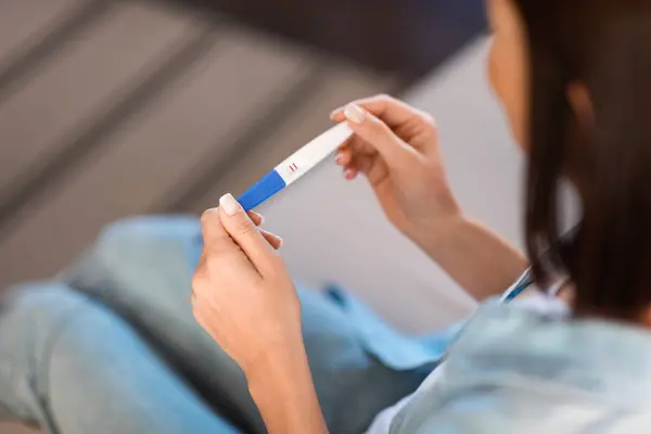 Unrecognizable young woman examines pregnancy positive test result, sitting on couch indoors. Concept of childbirth, fertility and pregnancy. Cropped, high angle shot, selective focus