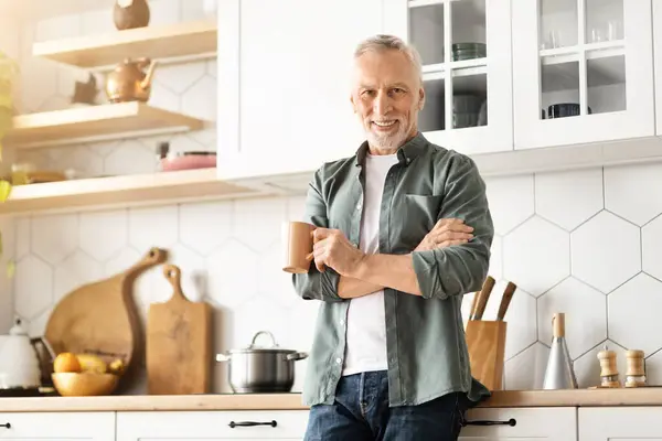Cheerful elderly gentleman standing with mug of coffee in hands, smiling senior man posing with arms crossed, enjoying happy morning in modern, well-equipped kitchen at home, free space
