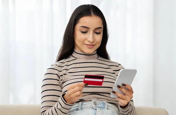 Positive young arab woman shopaholic, use credit card, mobile phone, pay for order, enjoy online shopping, check account app in living room interior. Money, finance, saving and profit, buy at home