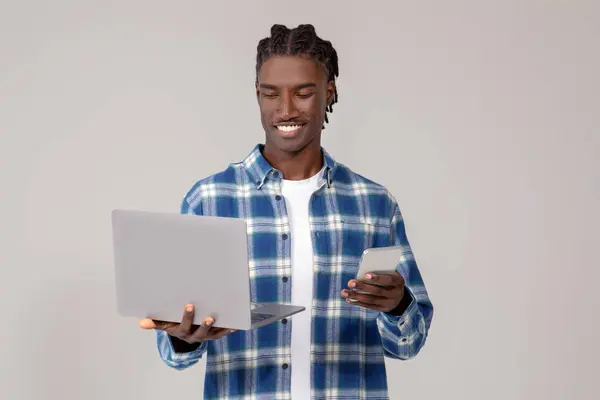 Laptop Kullanan Elinde Akıllı Telefon Tutan Odaklanmış Genç Siyahi Adam — Stok fotoğraf