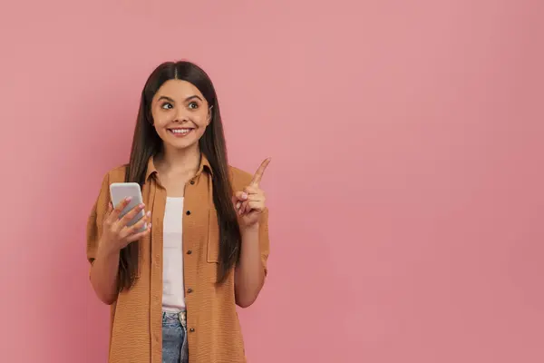 笑着拿着智能手机向上看的少女 快乐的少女 穿着粉色背景的休闲装 暗示着创新或灵感 — 图库照片