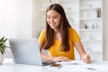 Çevrimiçi Ders. Gülümseyen Asyalı kadın dizüstü bilgisayar kullanıyor ve evinde otururken not alıyor, Kore 'li kadın uzaktan eğitimin ve uzaklığın tadını çıkarıyor, boş alan arıyor.