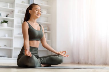 Güzel, genç Asyalı bir kadın evde yoga yaparken hafifçe gülümsüyor, sakin Koreli bir kadın oturma odasında minder üzerinde meditasyon yapıyor, Mudra yapıyor, rahatlama ve neşeyi somutlaştırıyor, boşluğu kopyalıyor.