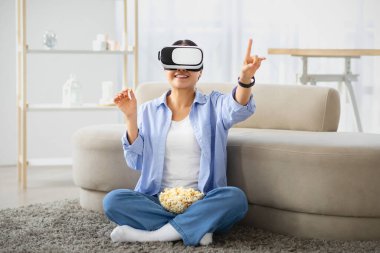 Heyecanlı Asyalı kadın evde VR kulaklık takıyor, rahat oturma odasında yerde oturuyor, efendim 'e dokunuyor, film izliyor, patlamış mısır yiyor, sanal gerçeklik deneyimi yaşıyor.
