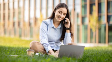 Gülümseyen profesyonel kadın, bilgisayarını açık havada çalışırken, modern iş hayatının esnekliğini kucaklayan, bir kahve fincanı yanında, akıllı telefonuyla sohbet etmekten hoşlanır.