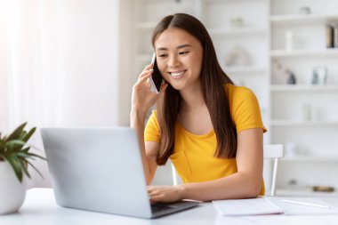 Genç Asyalı kadın cep telefonuyla konuşurken gülümsüyor ve dizüstü bilgisayarla çalışıyor. Güzel Koreli kadın, temiz, iyi organize edilmiş ofis alanında birden fazla görevi başarıyla yerine getiriyor.