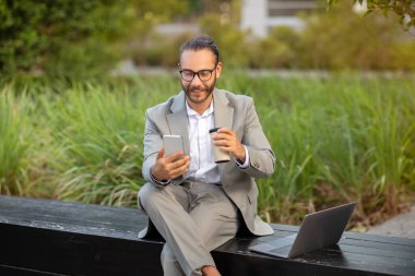 Parkta birden fazla iş adamı akıllı telefon ve kahveyle rahatlıyor, takım elbiseli gülümseyen erkek girişimci yanında dizüstü bilgisayarla bankta oturuyor, modern çalışma esnekliğini somutlaştırıyor.