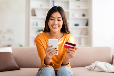 Evde kredi kartı ve akıllı telefon kullanan, internetteki mal ve hizmetler için ödeme yapan güzel Asyalı bir kadın. Mutlu Koreli bayan alışverişleri ya da bankacılık online, kopyalama alanı