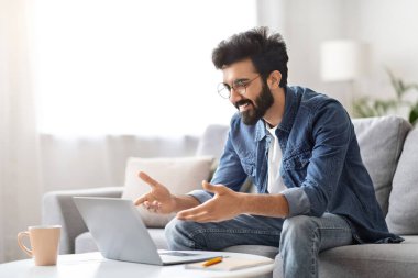 Yakışıklı Hintli adam dizüstü bilgisayarında video konuşması yapıyor, elleriyle işaret ediyor, rahat oturma odasındaki koltukta oturuyor, internet iletişiminin keyfini çıkarıyor, internetteki boşluğu kopyalıyor.