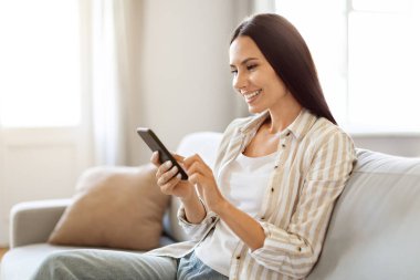 Evde akıllı telefonunu kullanan mutlu, rahat genç bir kadın. Gülümseyen milenyum kadını cep telefonu tutuyor, internetten alışveriş yapıyor ya da internette geziniyor, oturma odasında koltukta rahatça oturuyor, fotokopi çekiyor.