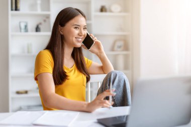 Sırıtkan Asyalı genç bir kadın dizüstü bilgisayarla masada oturuyor ve cep telefonuyla konuşuyor, mutlu Koreli serbest çalışan kadın ev ofisinde çalışırken cep telefonu konuşmasından hoşlanıyor, uzay boşluğunu kopyalıyor.