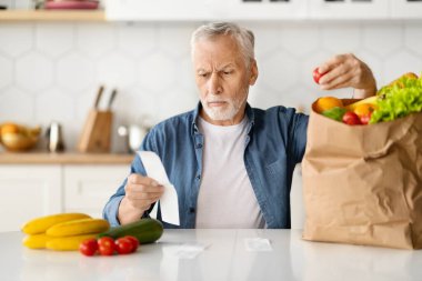Ciddi bir kıdemli adam faturaları kontrol ediyor ve yiyecek alışverişinden sonra bakkaliyeyi inceliyor. Mutfakta yaşlı ve düşünceli bir beyefendi oturuyor. Yaşlı bir beyefendi yüksek fiyatlardan rahatsız.
