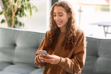 Mobil uygulamalar ve sosyal medya. Zoomer genç kız cep telefonuyla web sörfü yapıyor çevrimiçi boş zamanlarında, ev ortamında kanepede oturuyor. Gülümseyen genç bayan akıllı telefondan internette geziniyor.