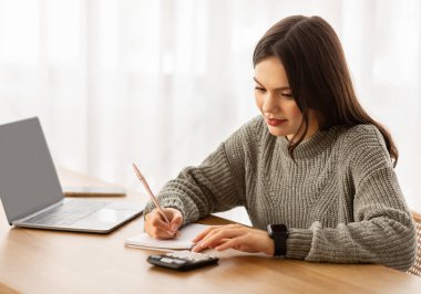 Aylık harcamaları sayan genç bir kadın, bütçe üzerinde çalışan, masa başında oturan, dizüstü bilgisayar ve hesap makinesi kullanan, not alan. Ofiste çalışan kadın finans müdürü, pazarlama raporu hazırlıyor.