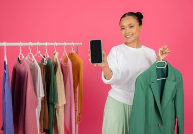 Mobil Alışveriş Uygulaması. Kamerada boş akıllı telefon gösteren neşeli Asyalı kadın giysi rafının yanında duruyor, moda ve tarz asistanı uygulaması öneriyor, pembe arka planda poz veriyor.