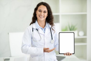 Neşeli Avrupalı bin yıllık kadın doktor elinde dijital tabletle beyaz ekran taklit alanı tutuyor, modern sağlık hizmetleri ve klinik içişleri teknolojileri için mobil uygulama öneriyor.