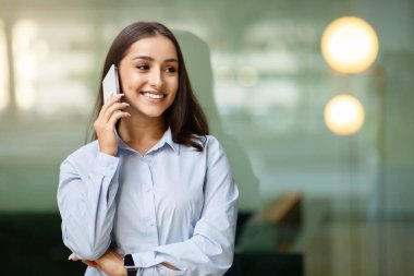 Zeki telefonuyla hoş bir sohbete girişen, kendine güvenen, genç, beyaz bir kadın. İnce bir gülümseme ve akıllı bir bilekle, kentsel bir ortamda.