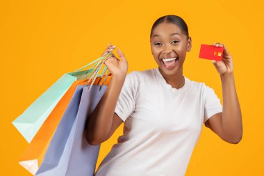 Kredi kartı ve renkli alışveriş torbaları tutan mutlu genç siyahi kadın, kameraya heyecanla gülümsüyor, reklam bankası ve perakende satış teklifi sarı arka planda. Satış sezonu
