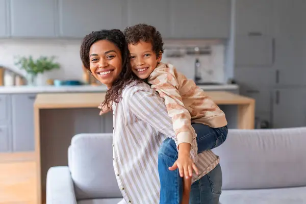 Cheerful black mother giving her happy son a piggyback ride at home, african american mom and preteen male kid smiling and enjoying playful moment together in cozy living room, copy space