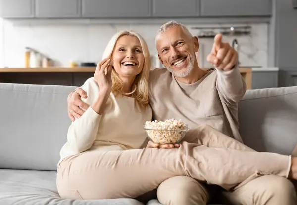 Happy senior couple watching TV together at home. Cheerful elderly man and woman sitting on couch, embracing, eating popcorn pointing at camera, enjoying movie, eating popcorn
