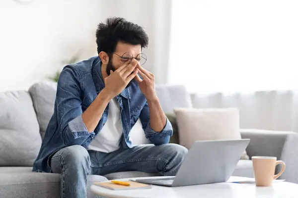 Tired Indian man rubbing his eyes, showing signs of fatigue or stress while working on laptop, exhausted eastern guy sitting on couch in living room, having eye-strain, copy space