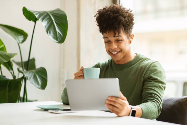 Happy millennial black guy with wireless earphones looking at tablet, surfing in internet, sitting at table with cup of coffee in cafe interior. Device, relax with music, funny video, have video call