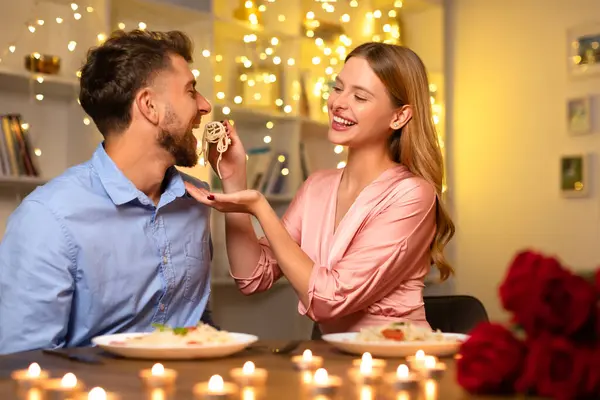 微笑的女人喂她的伴侣意大利面 两人在浪漫的气氛中 用蜡烛和仙女灯分享欢笑 营造出温暖的气氛 — 图库照片