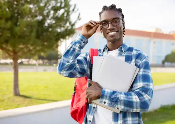 Smiling young black student guy in eyewear holding his laptop PC, engaged in e-learning, standing amid vibrant life of college campus outdoor, smiling to camera. Educational confidence