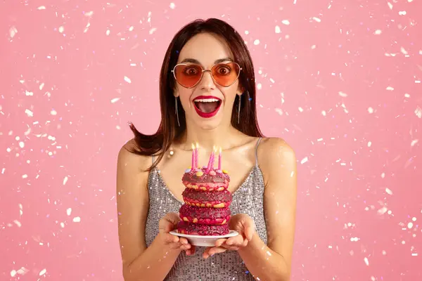 ピンクの背景にセッティのシャワーの中でキャンドルでカラフルなバースデーケーキを保持するサングラスと輝く銀のドレスで興奮した若い白人女性 クローズアップ — ストック写真
