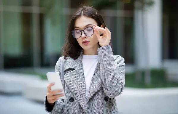 European young business lady looking at smartphone through eyeglasses, reading message and using mobile apps, standing on city street near modern business center building. Communication offer