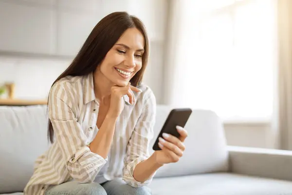 Χαμογελώντας Νεαρή Γυναίκα Χρησιμοποιώντας Smartphone Της Ενώ Χαλαρώνοντας Στον Καναπέ — Φωτογραφία Αρχείου