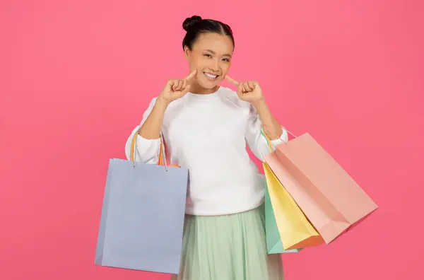 彼女のショッピングバッグを保持し 顔に触れる幸せなアジアの買い物客の女性 喜びを放射する陽気な韓国人女性 購入を楽しんでいるショップアホリック女性 ピンクのスタジオの背景に孤立 — ストック写真