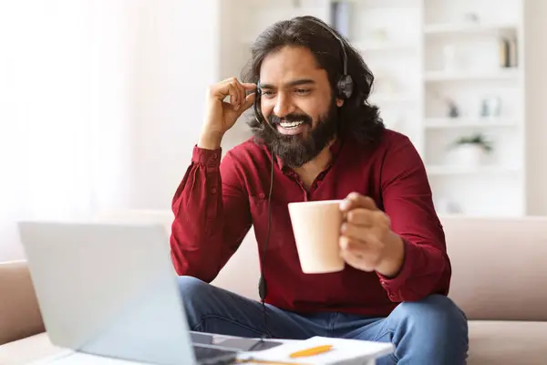 笑顔のミレニアル世代は 自宅からオンラインで学ぶ長髪の東部の男 彼の手でコーヒーマグとソファーに座って コンピュータとヘッドセットを使用して ウェビナーに出席 コピースペース — ストック写真