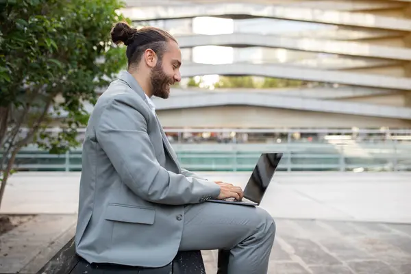 Contented modern businessman with topknot working on laptop outdoors, millennial male entrepreneur seated on bench in modern urban setting, enjoying remote work, typing on computer and smiling