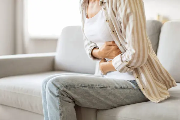 彼女の腹部を抱えている不快な若い女性は 十分に明るいリビングルームでソファーに座っている間 胃の痛みで気分が悪くなり 消化の問題を抱えている認識できない女性 クロップショット — ストック写真