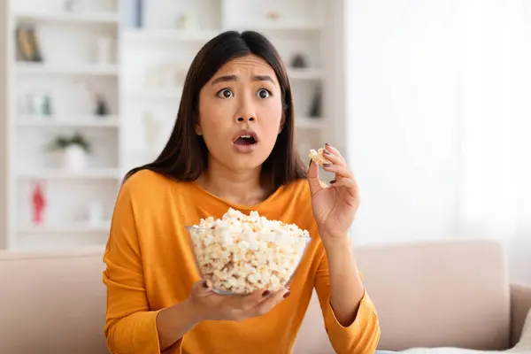 Closeup of frightened young asian woman watching scary movie at home and eating popcorn, scared millennial lady sitting on couch and looking at tv screen, emotionally reacting to shocked content