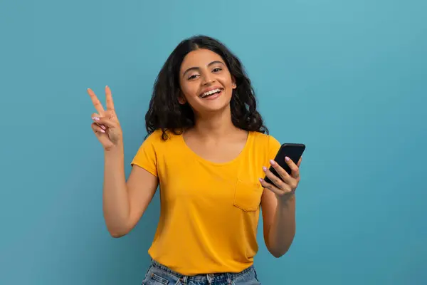 素晴らしいモバイルアプリ オンラインオファー ポジティブな魅力的なカーリー若いヒンドゥーの女性は 彼女の手で平和のジェスチャーと笑顔 青いスタジオの背景を示しています — ストック写真