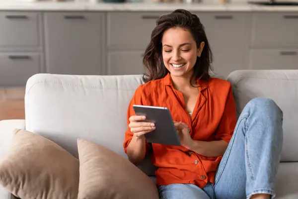 穿着鲜活橙色上衣的快乐的年轻女性沉浸在平板电脑中 舒舒服服地躺在家里时髦的灰色沙发上 自由自在 — 图库照片